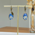 Boucles d'oreilles bronze Sajecréa, fleurs de Carotte sauvage sur fond bleu électrique.